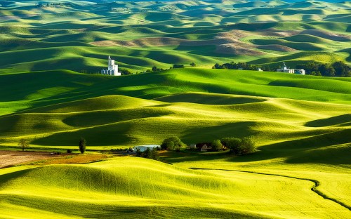 Lush green rolling hills with a small white church and farmhouses nestled among the landscape under a clear blue sky.