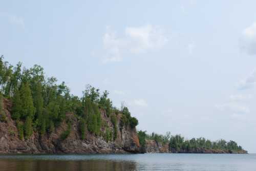 A serene view of rocky cliffs lined with greenery, reflecting in calm waters under a light blue sky.