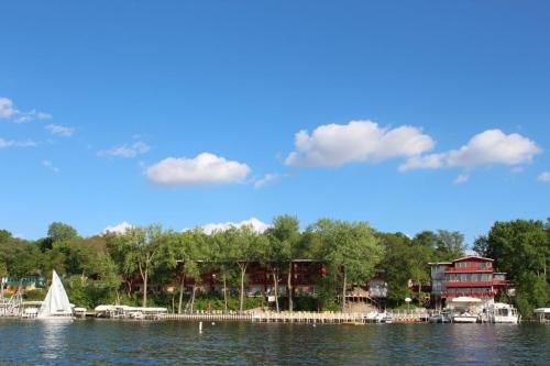 A serene lakeside view featuring a sailboat, lush trees, and colorful buildings under a clear blue sky with fluffy clouds.