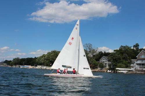 A sailboat with three people sails on a calm lake, surrounded by trees and houses under a partly cloudy sky.