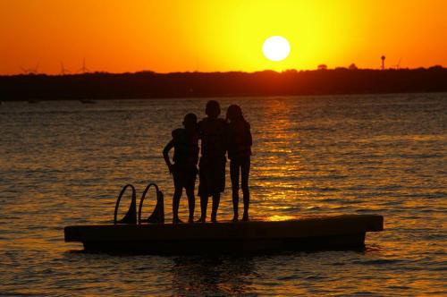 Silhouetted figures stand on a dock at sunset, with a vibrant orange sun setting over the water.