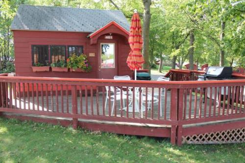 A small red cabin with a deck, surrounded by greenery, featuring a table and chairs, and an umbrella for shade.