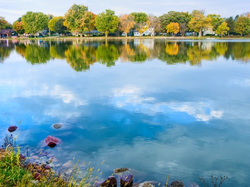 A serene lake reflecting colorful autumn trees and a clear sky, with rocks lining the shore.