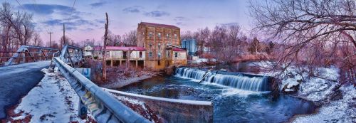 A panoramic view of a historic building by a river with a small waterfall, surrounded by bare trees and a winter sky.