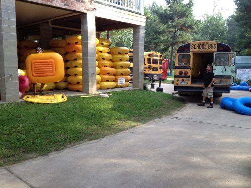 A scene with colorful inflatable rafts stacked near a school bus, with two people preparing for an outdoor activity.