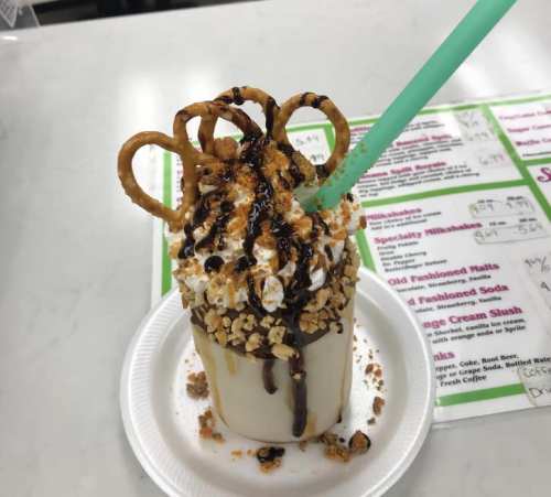 A decadent milkshake topped with whipped cream, chocolate drizzle, pretzels, and crushed nuts, served with a green straw.