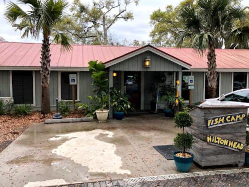 A rustic building with a red roof, palm trees, and potted plants, featuring a sign that reads "Fish Camp Hilton Head."