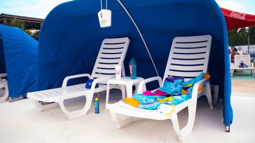 Two white lounge chairs under a blue canopy, with a colorful towel and drinks on the side.