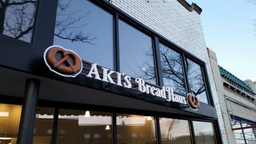 Sign for Aki's Bread Haus featuring a pretzel logo, displayed on a modern storefront with large windows.
