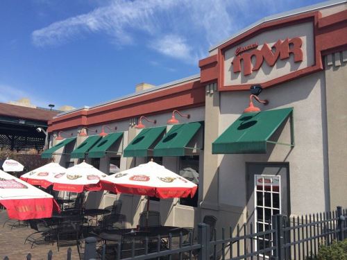 Exterior of a restaurant with green awnings, outdoor seating, and umbrellas on a sunny day.