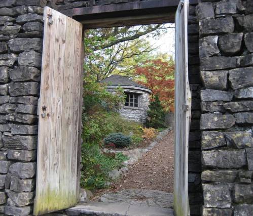 A stone doorway opens to a path leading to a round stone building surrounded by colorful foliage.