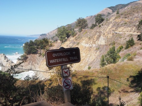Signpost indicating the direction to an overlook and Waterfall Trail, with a scenic coastal view in the background.