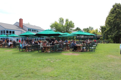 A lively outdoor dining area with green umbrellas, tables, and people enjoying meals on a sunny day.
