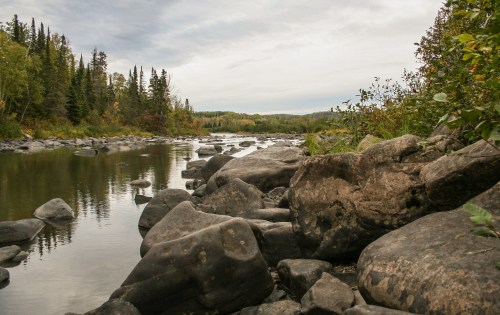 A serene river scene with smooth rocks along the shore, surrounded by trees and a cloudy sky.