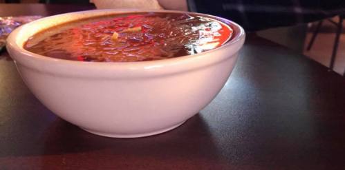 A white bowl filled with steaming soup, topped with shredded cheese, sitting on a dark table.