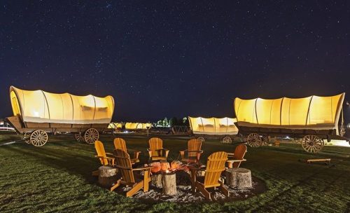 A starry night sky over illuminated covered wagons and a circle of wooden chairs around a campfire.
