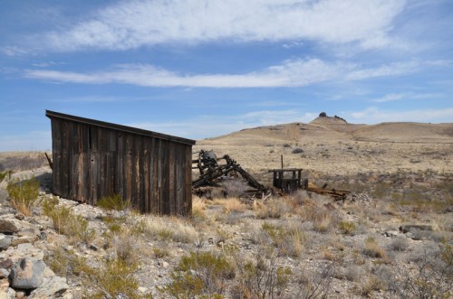 A weathered wooden structure sits in a barren landscape, with rocky hills and sparse vegetation under a blue sky.