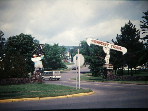 Signage for the Gunflint Trail, featuring a bear and a large arrow, with a van parked nearby on a scenic road.