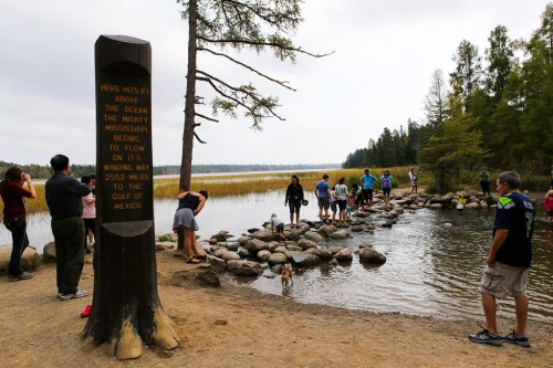 Visitors gather near a marker indicating the start of the Mississippi River, with a rocky path and water in the background.