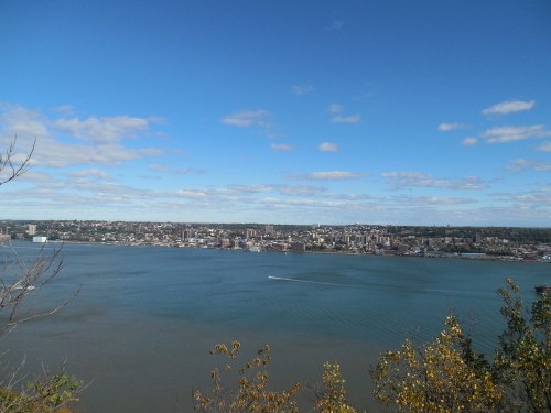 A panoramic view of a city skyline along a river under a clear blue sky with scattered clouds.