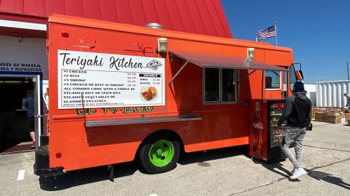 A vibrant orange food truck named "Teriyaki Kitchen" with a menu displayed, parked near a red building.