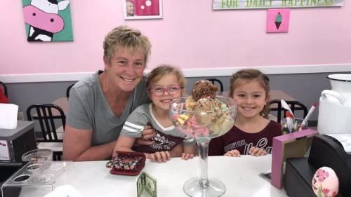 A smiling woman and two girls pose with a large ice cream sundae at a colorful ice cream shop.