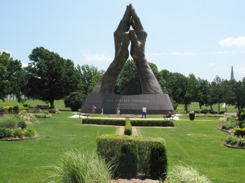 A large sculpture of two hands in prayer at Oral Roberts University, surrounded by green grass and trees.