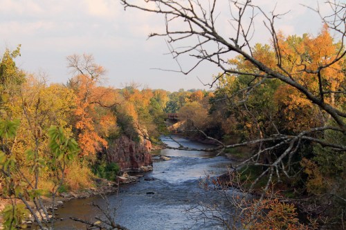 A serene river flows through a landscape of autumn trees, with rocky banks and a bridge in the distance.