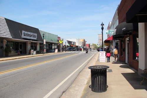 A quiet street lined with shops, a motorcycle passing by, and pedestrians walking on a sunny day.