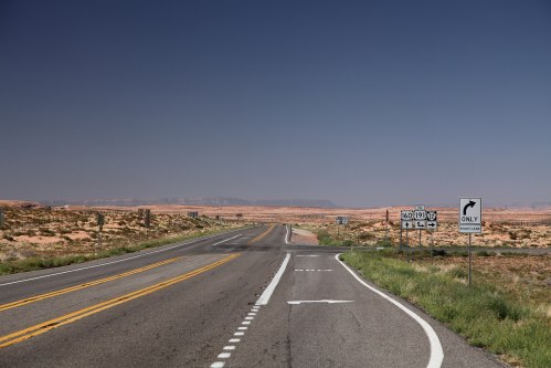 A wide, empty road stretches into the desert, with directional signs and a clear blue sky above.