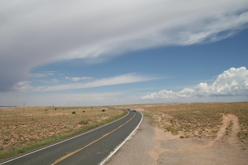 A winding road stretches through a vast, arid landscape under a blue sky with scattered clouds.