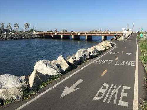 A bike lane along a waterfront with a bridge in the background and large rocks lining the path. Clear blue sky above.
