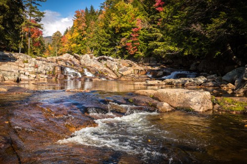 A serene river flows over rocks, surrounded by colorful autumn trees under a clear blue sky.