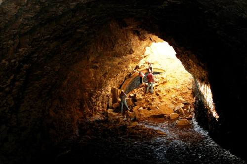 Two people explore a rocky cave, with sunlight illuminating the entrance and water at the base.