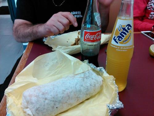 A burrito wrapped in yellow paper, with a Coca-Cola and Fanta Pineapple bottle on a table. Hands are visible in the background.