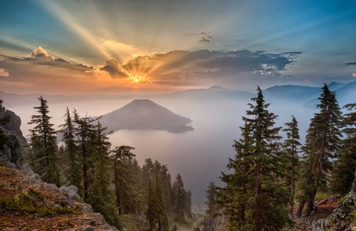 Sunset over a misty lake surrounded by mountains and evergreen trees, with rays of light breaking through the clouds.