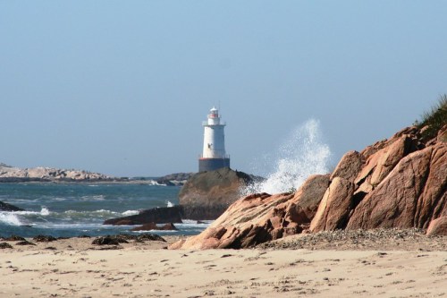 A lighthouse stands on a rocky shore, with waves crashing against the rocks and a clear blue sky in the background.