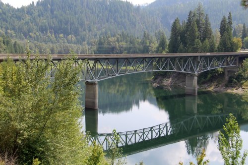 A metal bridge spans a calm river, surrounded by lush green trees and mountains in the background.
