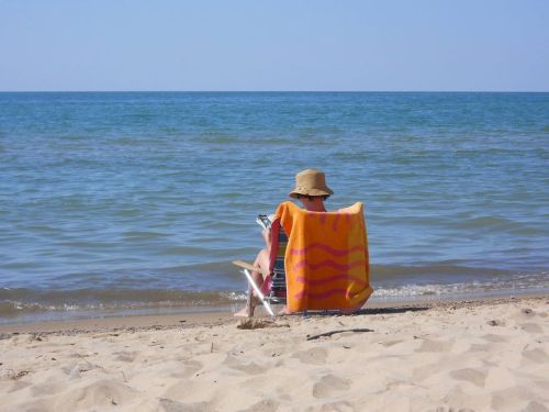 A person sitting on a beach chair, reading a book, with a towel draped over their shoulders and the ocean in the background.
