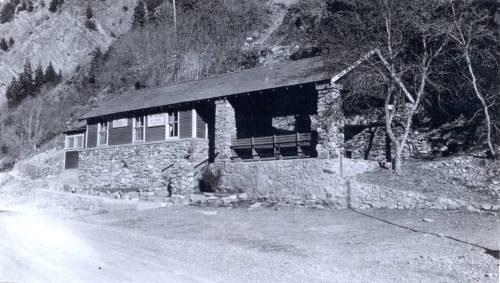 Historic stone building with a wooden porch, set against a mountainous landscape. Black and white photo.