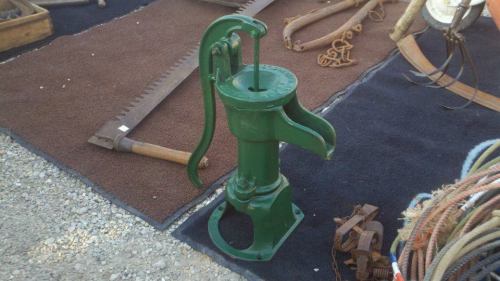 A vintage green hand pump stands on a dark surface, surrounded by various antique tools and equipment.