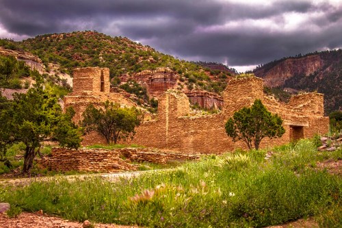 Ancient stone ruins surrounded by greenery and mountains under a cloudy sky.