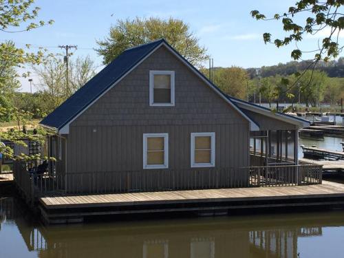 A small, wooden house on a dock by a calm body of water, surrounded by trees and a marina in the background.