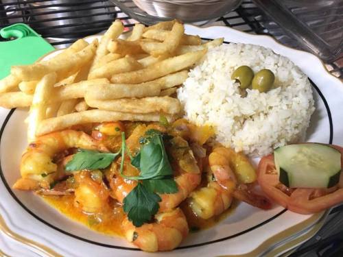 A plate of shrimp in sauce with rice, fries, olives, and fresh cucumber and tomato slices, garnished with parsley.