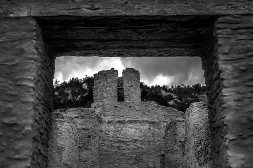 Black and white image of ancient stone ruins framed by wooden beams, with a cloudy sky in the background.