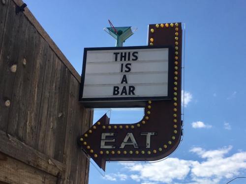 Sign with lights reading "THIS IS A BAR" with an arrow pointing to "EAT," set against a blue sky.