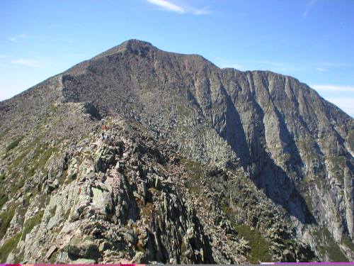 A rugged mountain peak with rocky terrain and hikers on a narrow ridge under a clear blue sky.