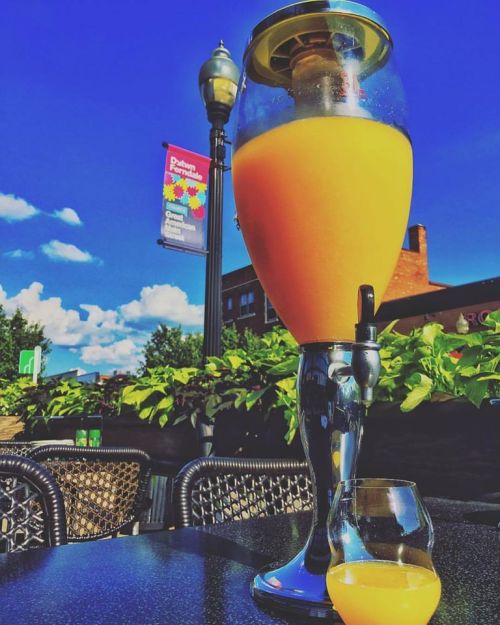 A large glass of orange drink with a tap, next to a smaller glass, set against a blue sky and greenery.