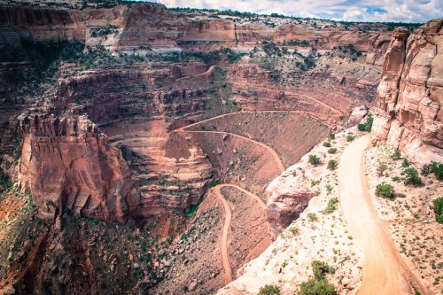 A winding dirt road curves through a deep, rocky canyon under a partly cloudy sky.