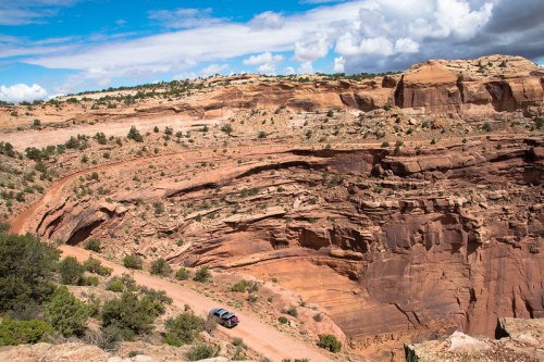A winding dirt road curves around a rocky canyon landscape under a partly cloudy sky. A car is visible on the road.